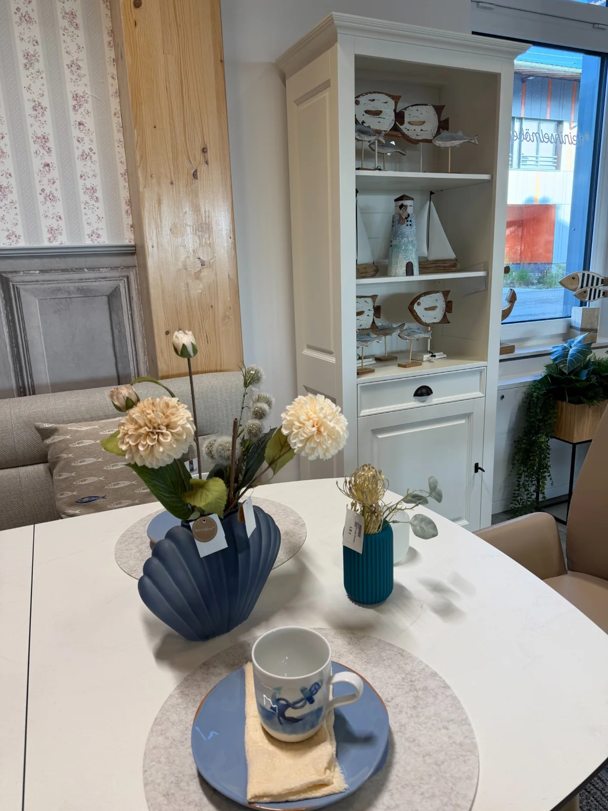Maritime Tischdekoration mit blauer Muschelvase, Tasse und weißer Vitrine mit Fischfiguren